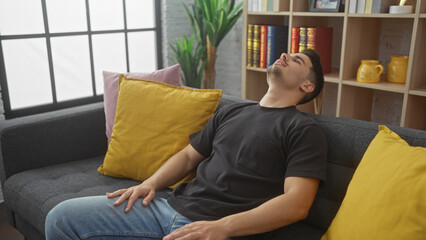 Relaxed hispanic man napping on couch at home with bookshelf and decorative pillows in the...