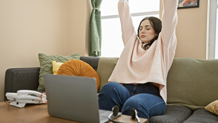 A young woman stretching on a couch in a living room with a laptop, showing a casual home working...