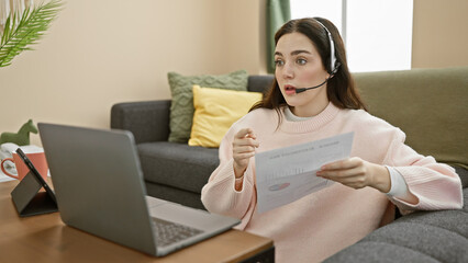 A young caucasian woman is working from home, conducting a video call while holding a document,...