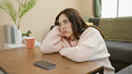 Pensive young woman in pink sweater resting chin on hands at home with coffee and smartphone on...