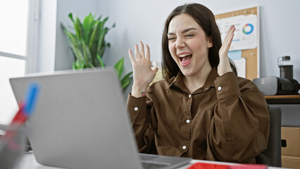 A delighted young caucasian woman is exuberantly expressing joy at her office workplace, providing...