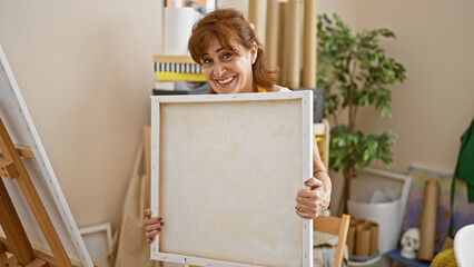 Mature redhead woman smiling and holding a blank canvas in a bright art studio.