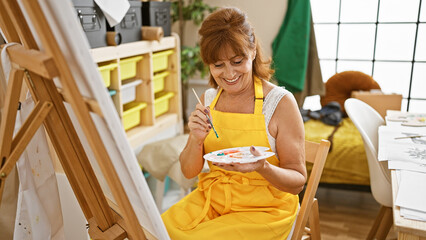 Smiling mature woman painting in a bright art studio, wearing a yellow apron