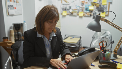 Mature woman using laptop in cluttered office space, indicative of investigative work.