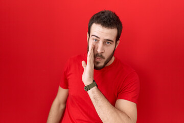 Young hispanic man wearing casual red t shirt hand on mouth telling secret rumor, whispering...