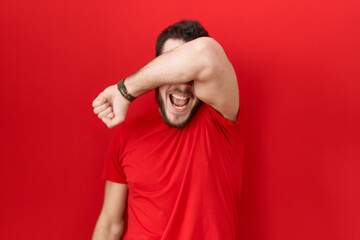 Young hispanic man wearing casual red t shirt covering eyes with arm smiling cheerful and funny. blind concept.