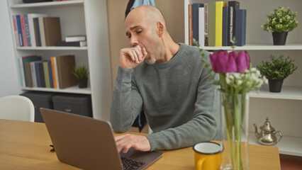 A bald, bearded, young hispanic man in a gray sweater yawning while using a laptop in a cozy home...