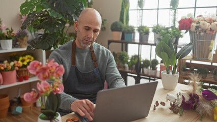 A bald man works at his laptop in a vibrant flower shop filled with various plants and flowers.