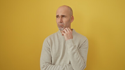 A handsome bald man with a beard, wearing a sweater, ponders thoughtfully against a yellow isolated...