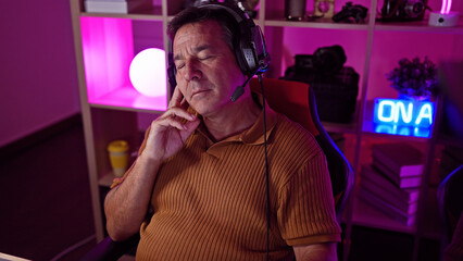A thoughtful mature man wearing a headset sits in a dimly lit gaming room with neon lights at night, reflecting modern home entertainment.