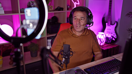 A mature man with headphones sits in a colorful gaming room at night, speaking into a microphone...