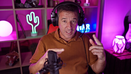 Mature man in headphones streaming in a neon-lit gaming room, pointing at phone.
