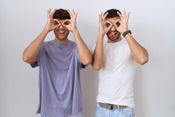 Homosexual gay couple standing over white background doing ok gesture like binoculars sticking...