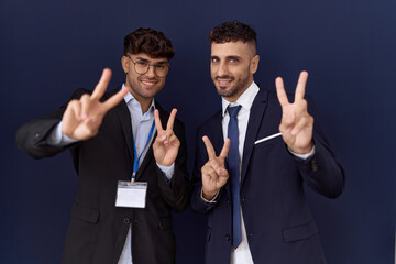 Two hispanic business men wearing business clothes smiling looking to the camera showing fingers doing victory sign. number two.