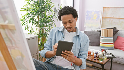 A thoughtful young man with curly hair browsing a tablet in a creative indoor studio setting surrounded by art supplies.