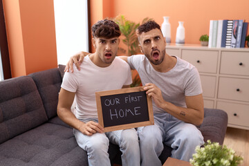 Homosexual gay couple holding blackboard with first home text in shock face, looking skeptical and...