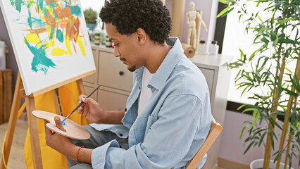 Young adult man with curly hair painting on canvas in art studio, showcasing creativity and...