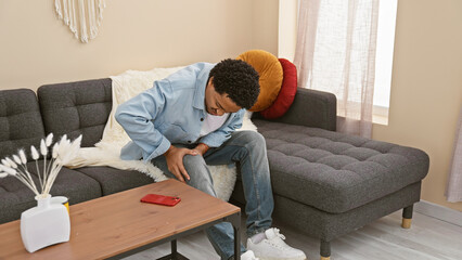 A young adult man with curly hair experiencing knee pain while seated on a couch indoors.