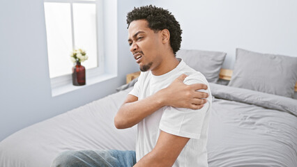 A young man expressing discomfort while holding his shoulder in a modern bedroom setting.