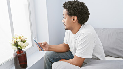 African american man with curly hair using smartphone in modern living room, sitting on a couch by the window.