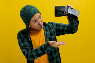 Dismayed Asian man stares at his empty wallet. Isolated on a yellow background. Financial hardship,...
