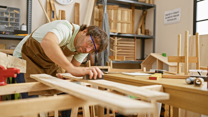 A young bearded man sands wood in an indoor carpentry workshop, wearing safety glasses and a brown...