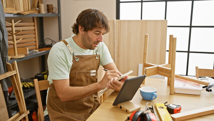 Bearded man attentively using tablet in a well-equipped carpentry workshop.