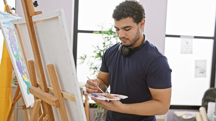 A young artist man with headphones paints on a canvas in an indoor studio setting.