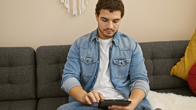 A young man with beard in casual denim jacket focused on using a tablet while sitting on a grey sofa indoors. - Powered by Adobe
