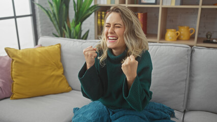 A cheerful young woman celebrates a joyful moment alone on a gray couch in a cozy living room...