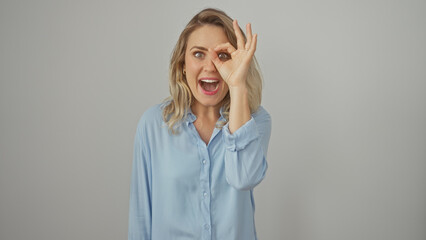Smiling young blonde woman in blue shirt making okay sign with hand over eye on white background