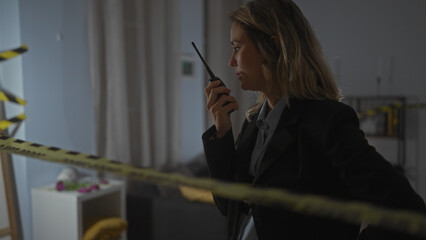 A focused blonde woman wearing a black suit communicates on a handheld radio at an indoor crime...