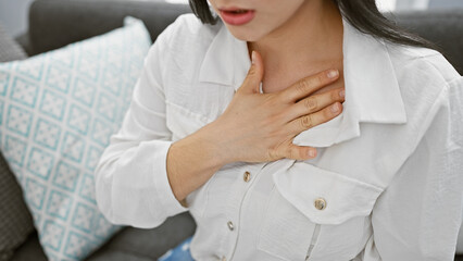 A young hispanic woman in casual attire is indoors, touching her chest, displaying a subtle...