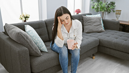 Hispanic woman holding coffee mug appearing pensive in a modern living room setting.