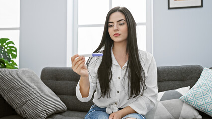 A young hispanic woman intently looks at a pregnancy test while sitting in her modern living room.