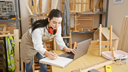 Hispanic woman planning carpentry work in a workshop using a laptop and notepad