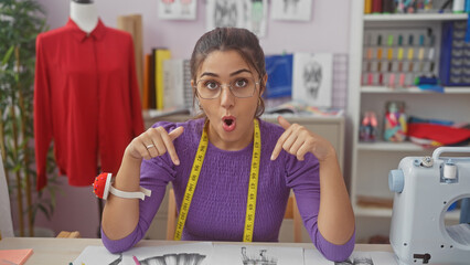 Surprised hispanic woman with glasses in a tailor shop, pointing at dress designs, surrounded by sewing equipment.