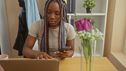 African american woman with braids using a phone at home, sitting by a vase of flowers in an indoor...