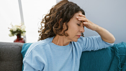 A stressed mature hispanic woman with curly hair touches her forehead indoors, expressing worry or...