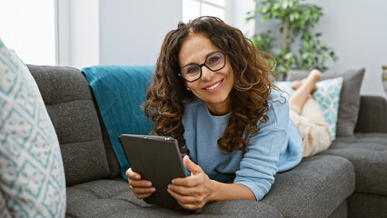 A smiling mature hispanic woman with curly hair, lying comfortably on a couch at home, holding a...