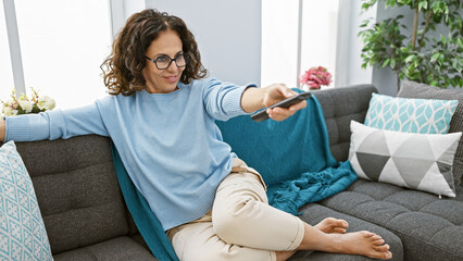 A middle-aged woman with curly hair comfortably relaxes at home, extending a remote control while...