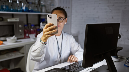 A mature woman scientist in a laboratory examines a smartphone while working at a computer,...