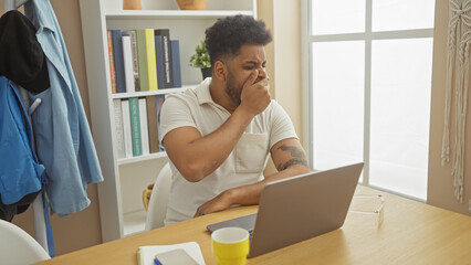 An adult african american man looks tired while working from home in a well-organized living room.