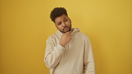 A thoughtful african american man in a hoodie poses against a yellow background.