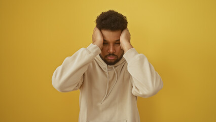 Exhausted african american man in beige hoodie with hands on head against a yellow background.