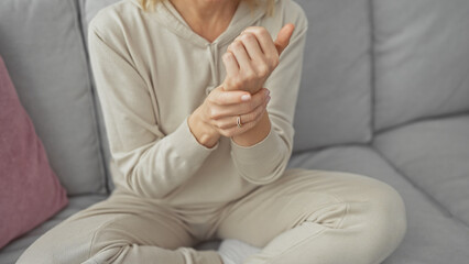 A young woman in casual wear sits indoors on a sofa, holding her wrist as if in pain or discomfort.