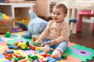 Adorable toddler playing with wooden construction blocks sitting on floor at kindergarten