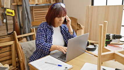 Mature woman multitasking on phone and laptop in a carpentry workshop.