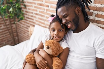 Father and daughter hugging each other sitting on bed holding teddy bear at bedroom
