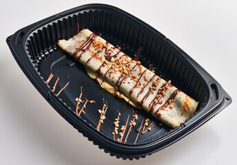 Delicious crepes with chocolate and nuts served in a black takeout container against a white...